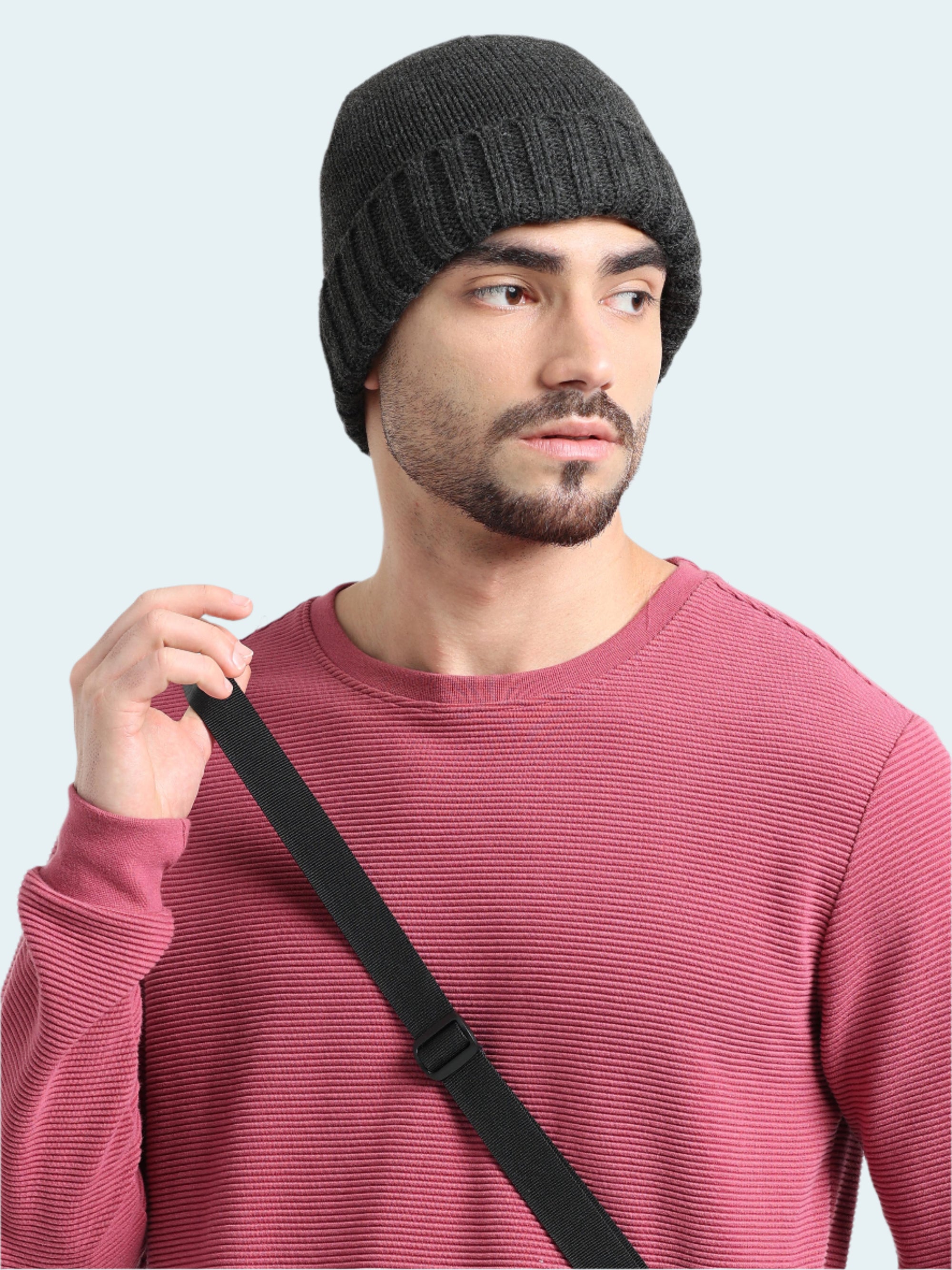 Man wearing a black knit beanie and pink sweater with a black strap over his shoulder on a white background