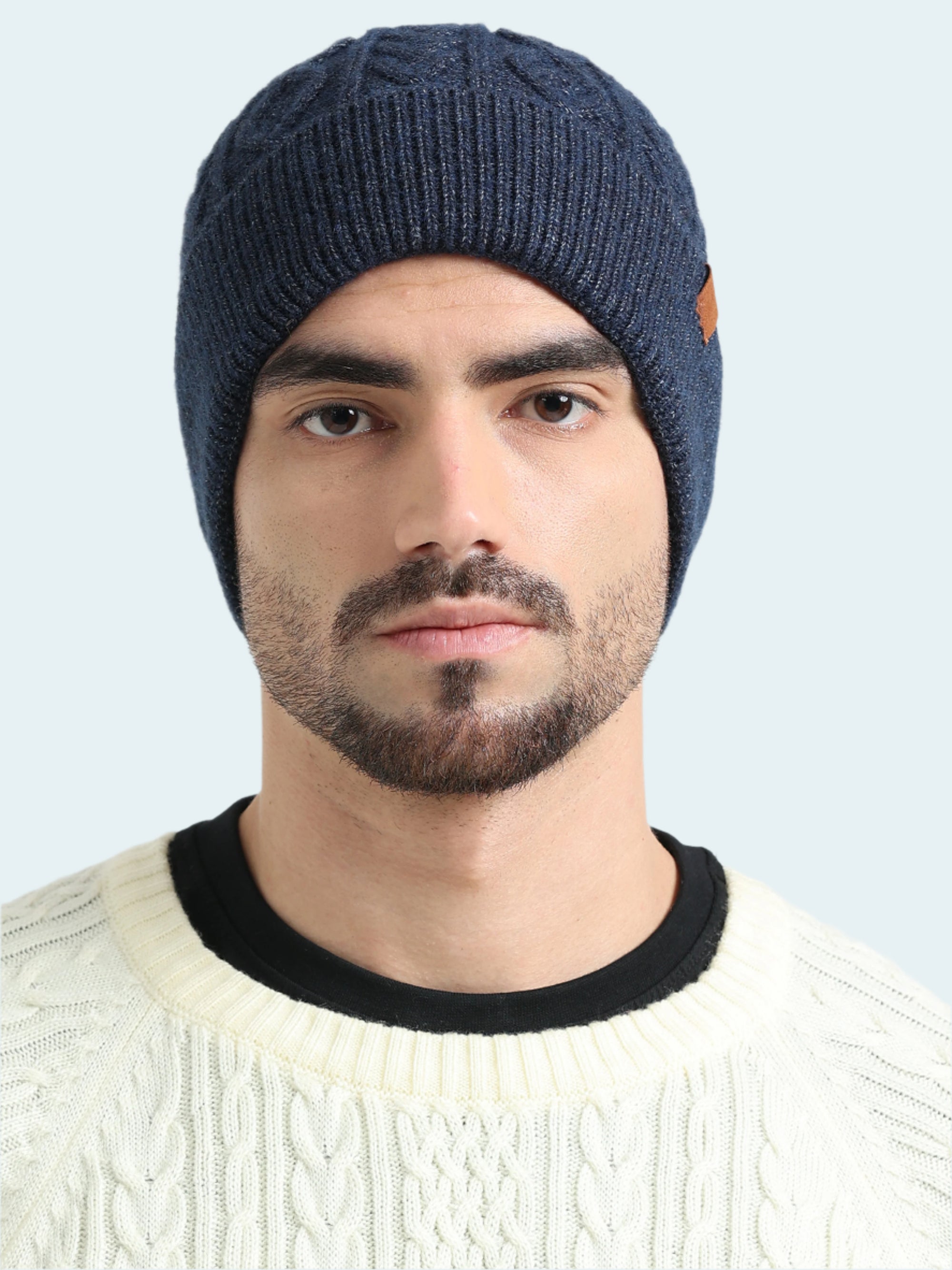 Man wearing a blue knitted beanie and white sweater with black collar against a light gray background