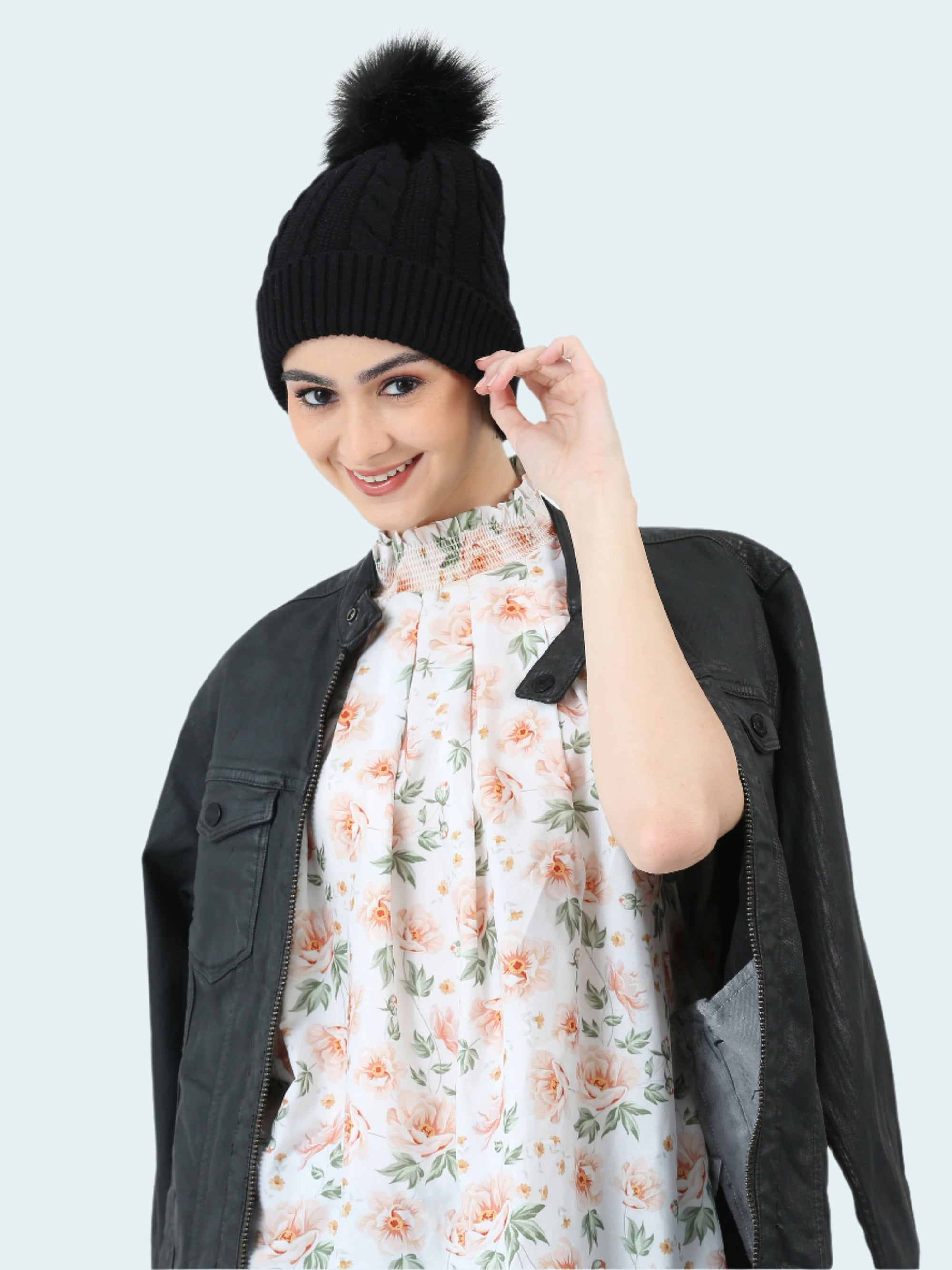 Woman wearing a black knit beanie with a pom-pom, floral scarf, and dark jacket on a white background