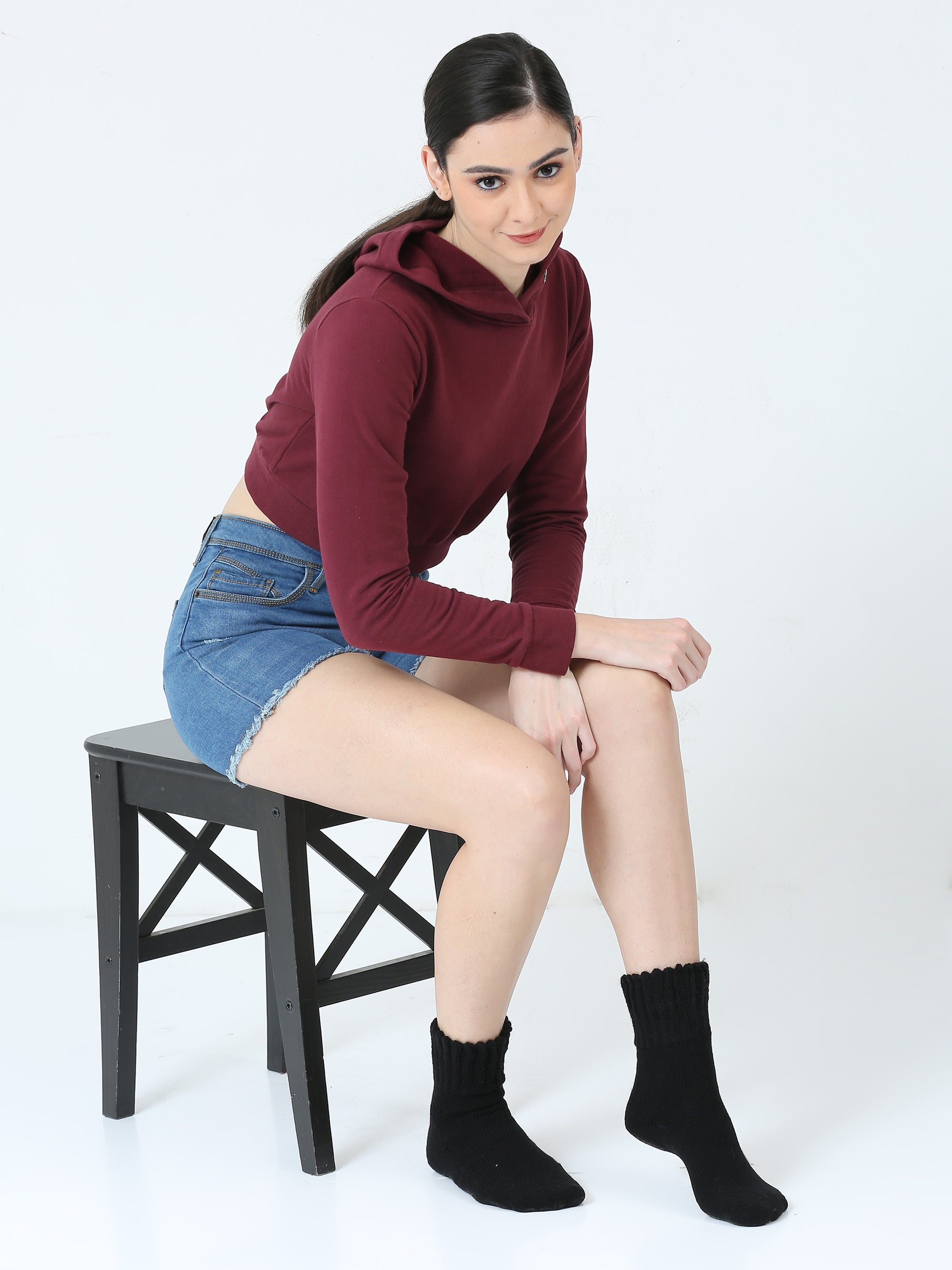 Woman sitting on a stool wearing a maroon hoodie, denim shorts, and black socks.