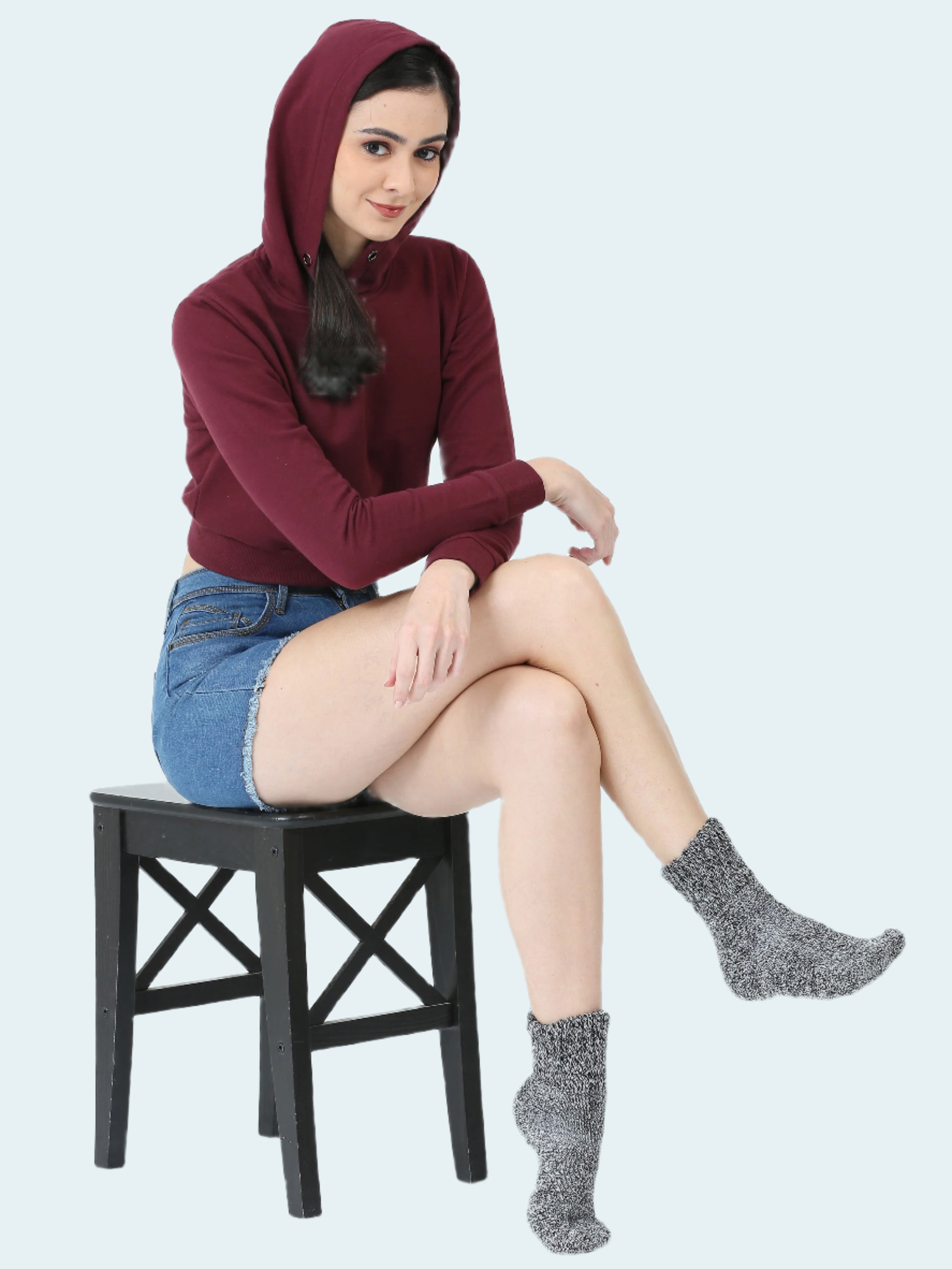 Woman wearing a maroon hoodie, denim skirt, and gray socks sitting on a black stool against a white background.