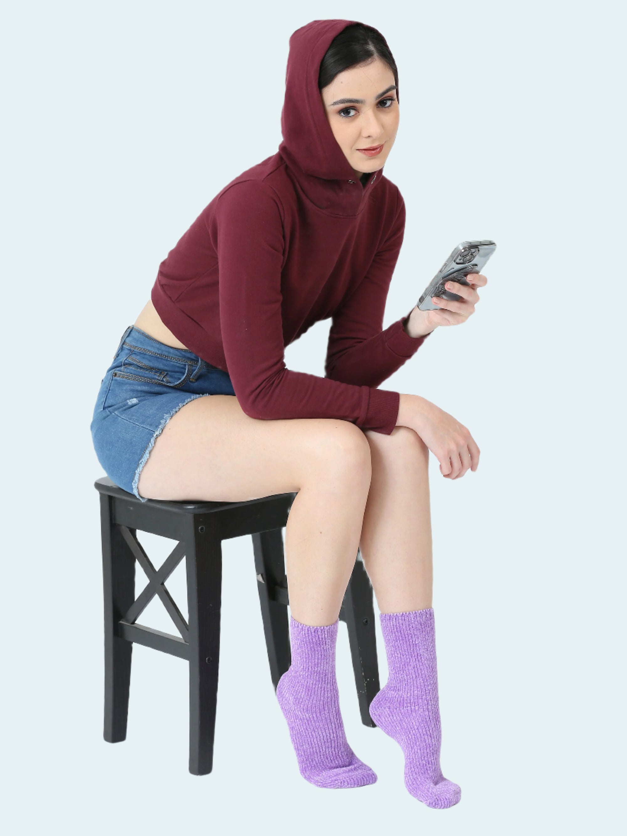 Woman sitting on a chair wearing a maroon hoodie, denim shorts, and purple socks, holding a phone.