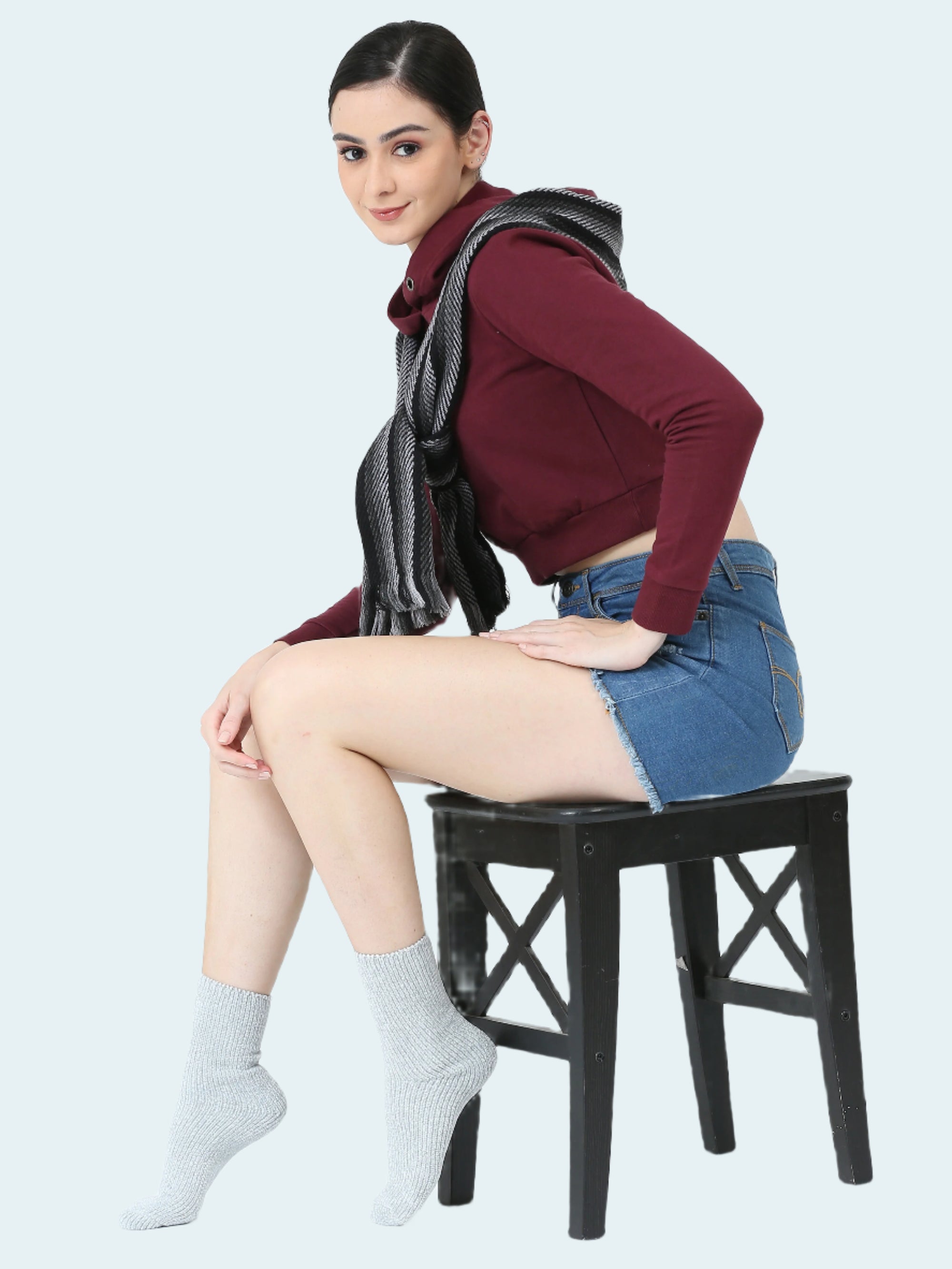 Woman sitting on a black chair wearing a maroon top, denim skirt, and gray socks against a white background