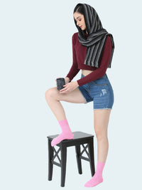 Woman wearing a burgundy top, denim shorts, pink socks, and a gray hijab sitting on a black stool.