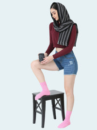 Woman wearing a burgundy top, denim shorts, pink socks, and a gray hijab sitting on a black stool.