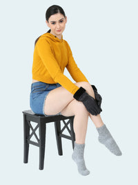 Woman sitting on a black stool wearing a yellow hoodie, denim shorts, gray socks, and black gloves on a white background.