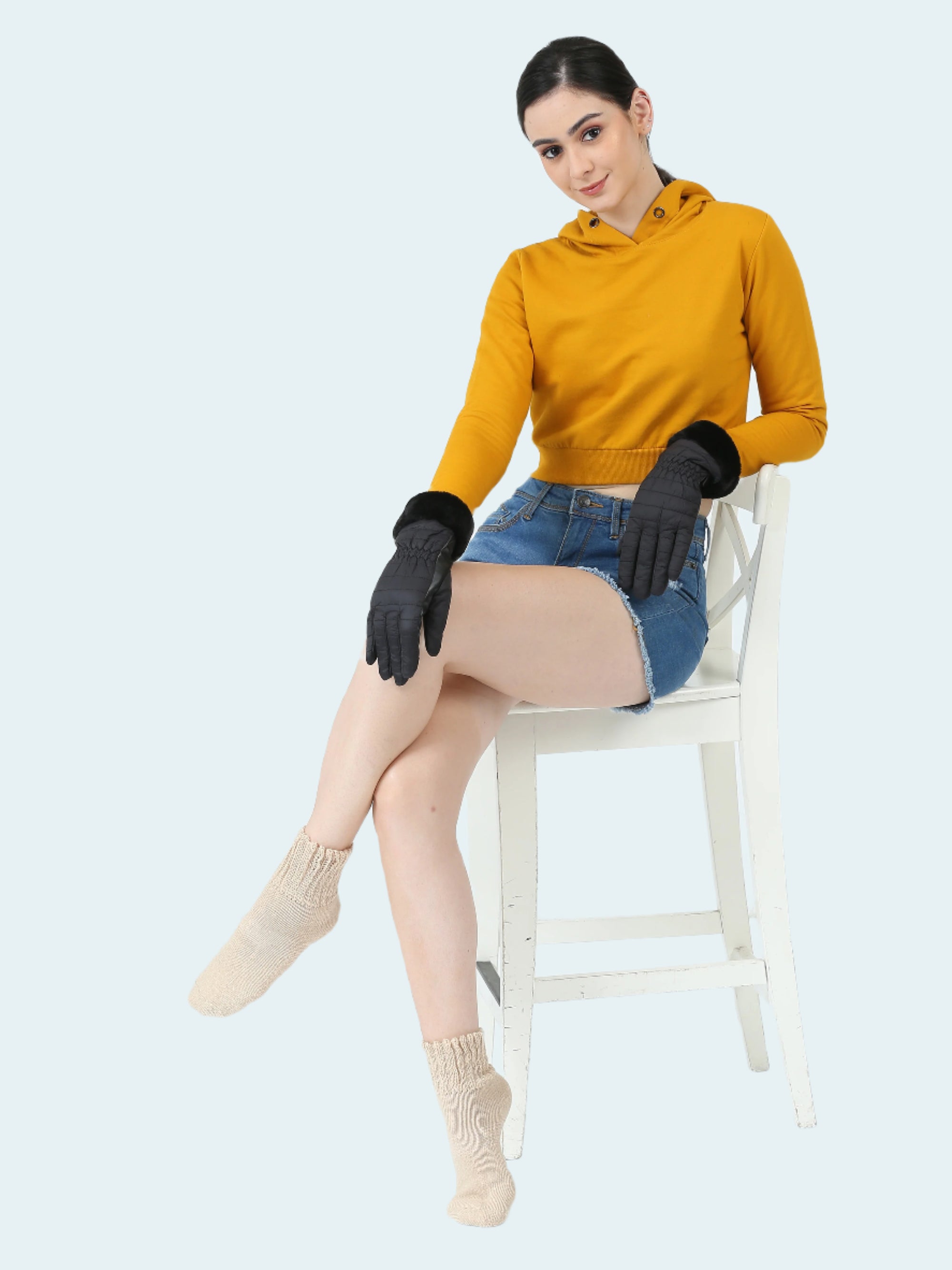Woman wearing a mustard yellow hoodie, denim skirt, and black gloves sitting on a white chair.