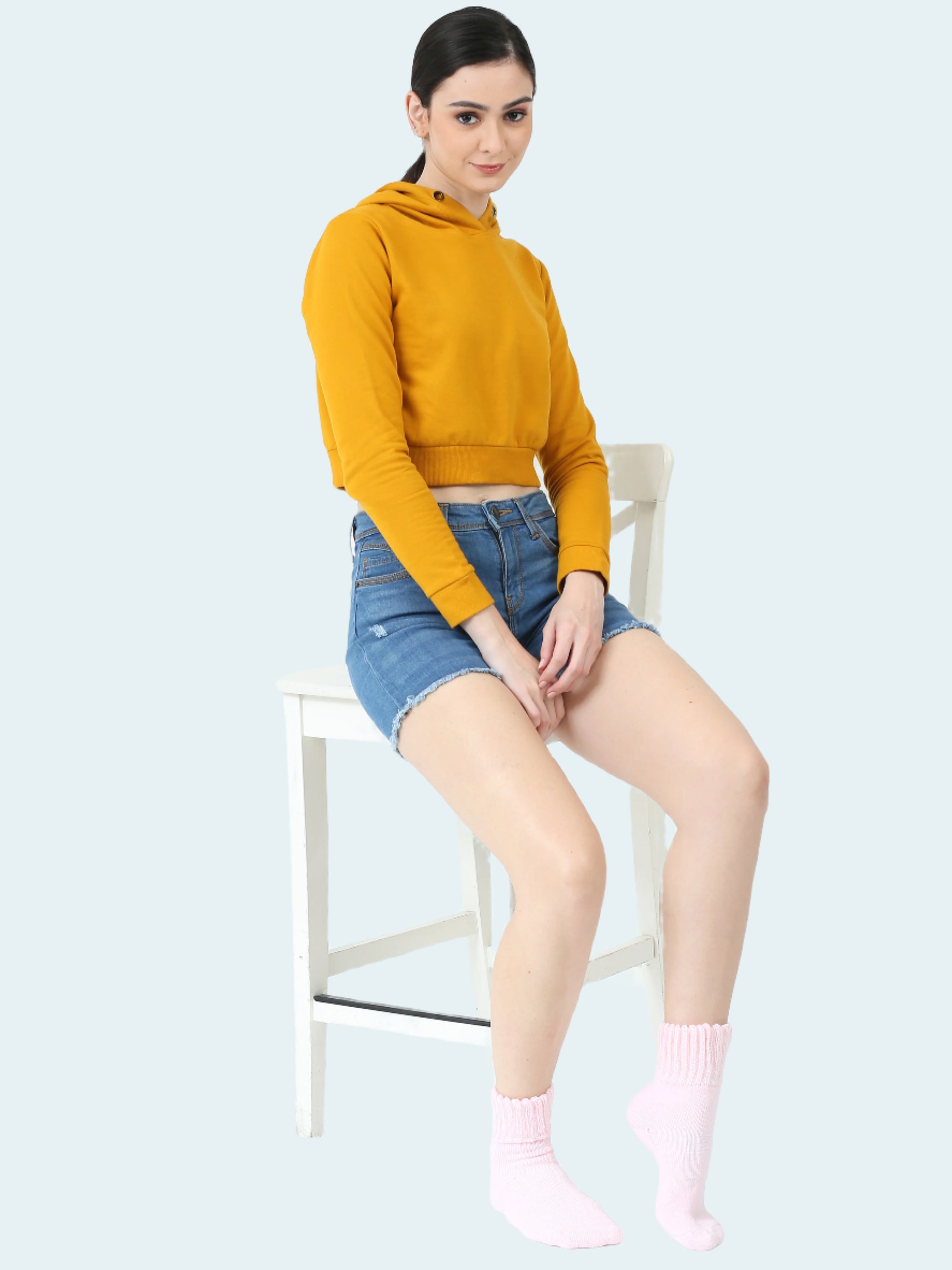 Woman wearing a mustard yellow hoodie, denim shorts and pink socks sitting on a chair.