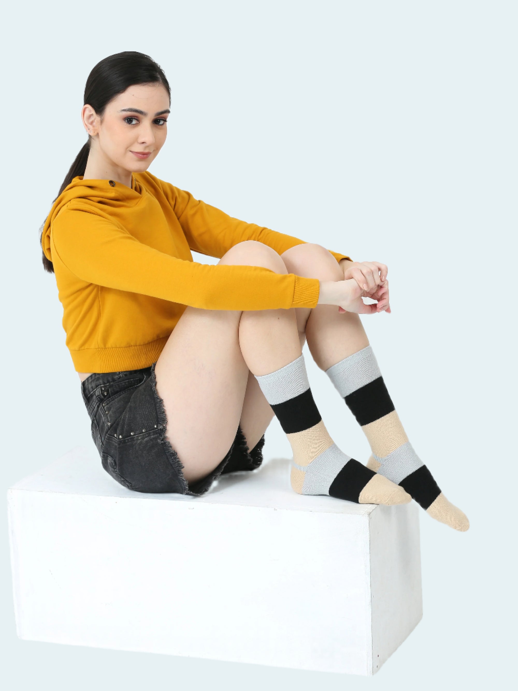 Woman wearing mustard yellow hoodie and black and gray striped socks sitting on a white box against a white background