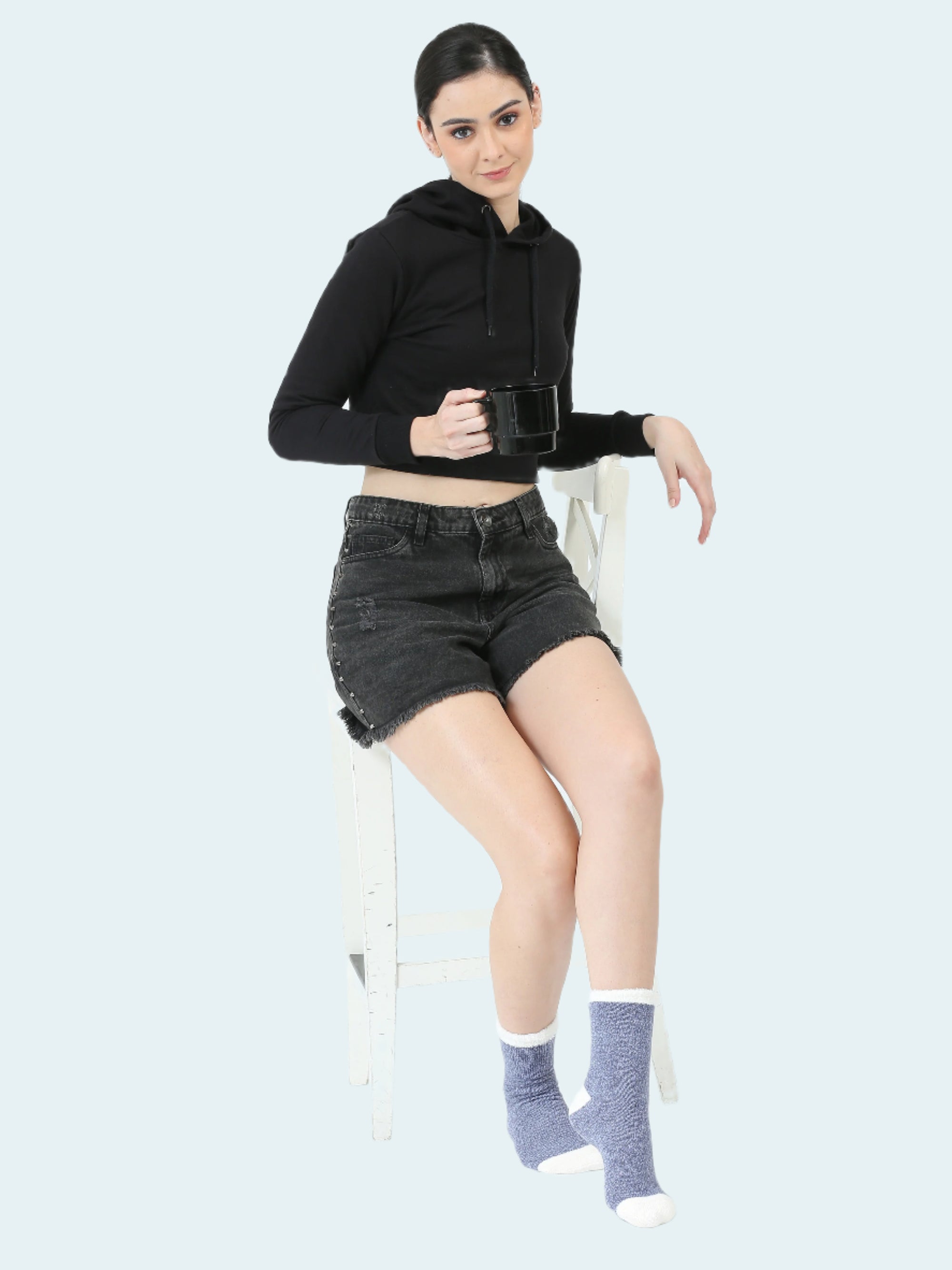 Woman sitting on a chair wearing a black hoodie, denim shorts, and gray socks.