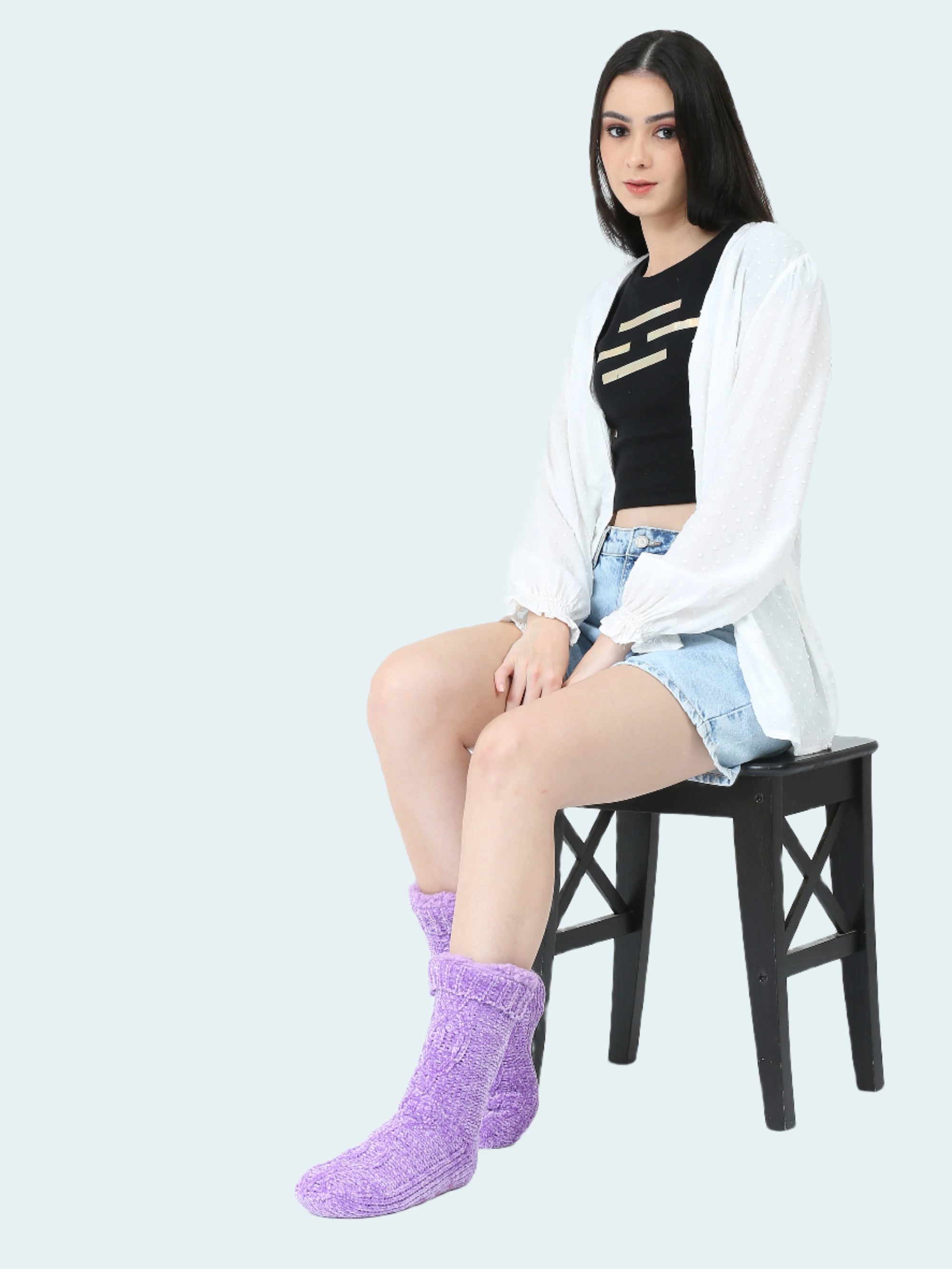 Woman sitting on a chair wearing purple socks, black top, and light blue shorts with a white cardigan.