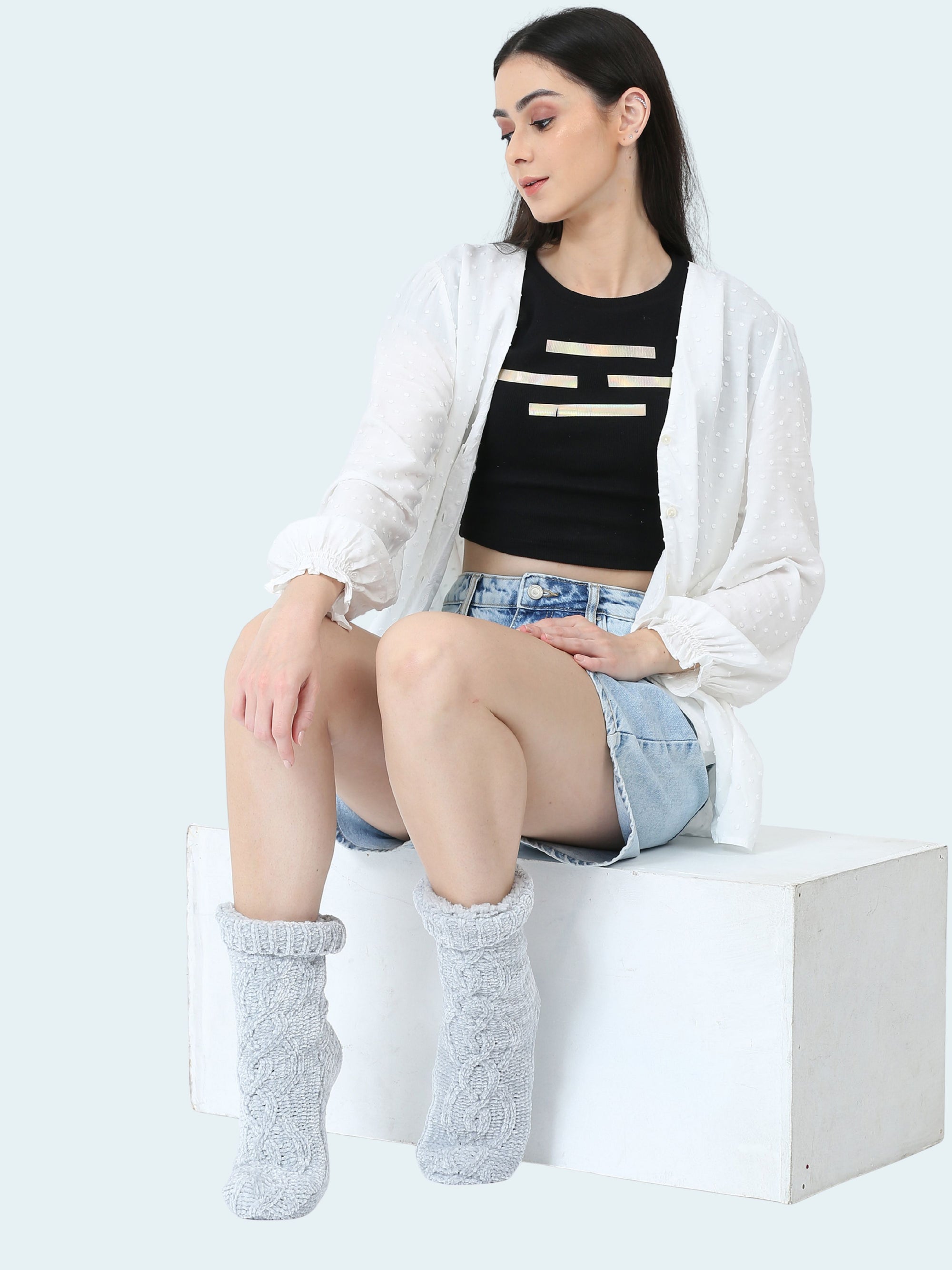 Woman sitting on a white surface wearing gray socks, a black crop top, denim shorts, and a white cardigan.