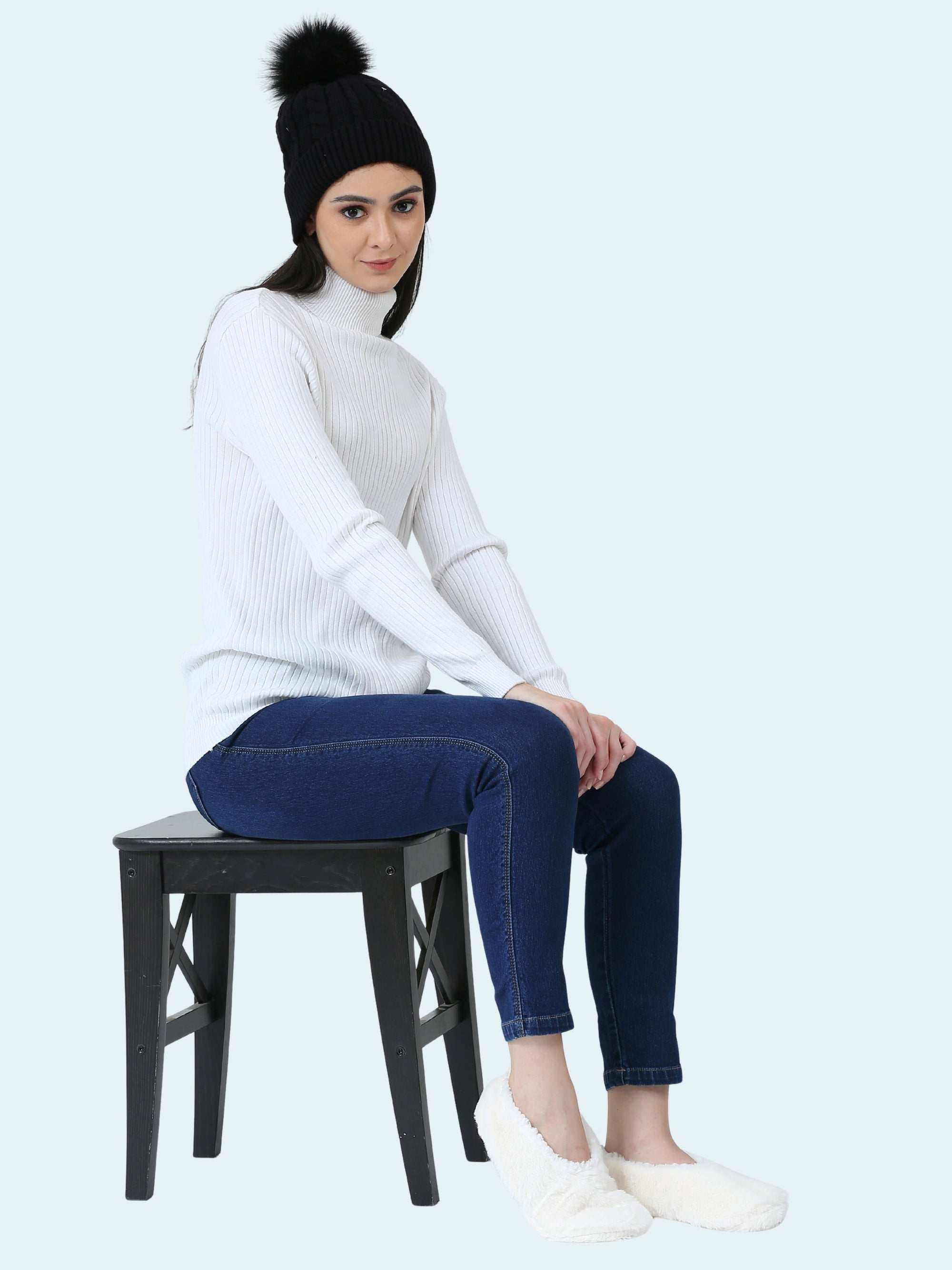 Person sitting on a stool wearing a white sweater, dark jeans, and a black beanie with a pom-pom and white slipper socks