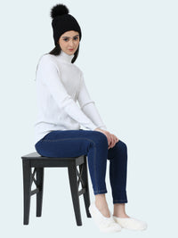 Person sitting on a stool wearing a white sweater, dark jeans, and a black beanie with a pom-pom and white slipper socks