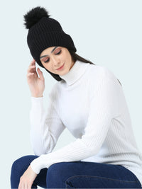 Woman wearing a black knit beanie with a pom-pom, white turtleneck sweater, and blue jeans on a light gray background.