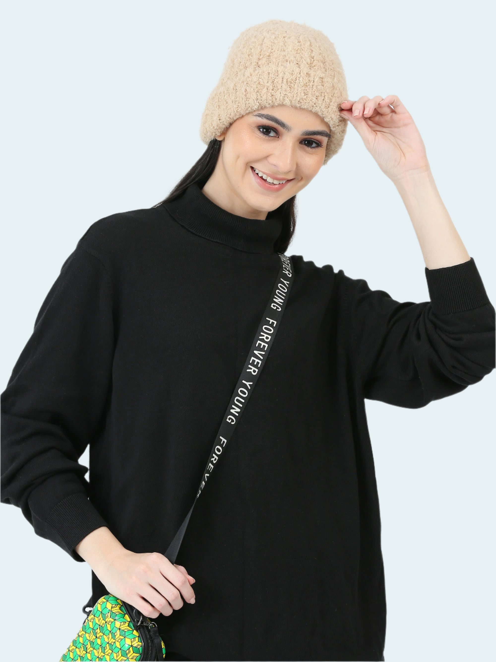 Woman wearing a black turtleneck sweater with text, beige beanie, and holding a colorful bag on a white background