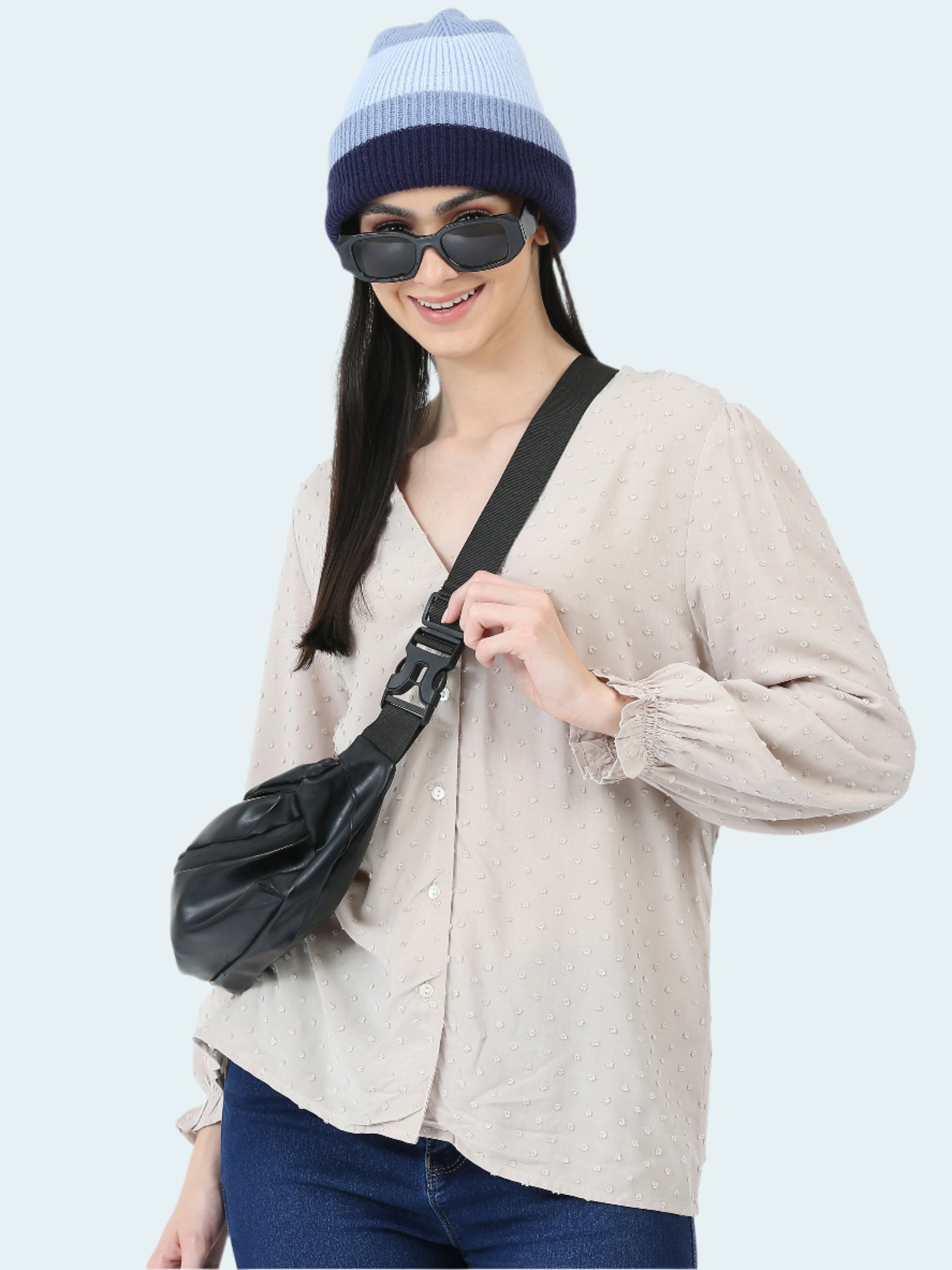 Woman wearing a beige shirt, blue jeans, and a two-tone beanie with sunglasses, holding a black bag.
