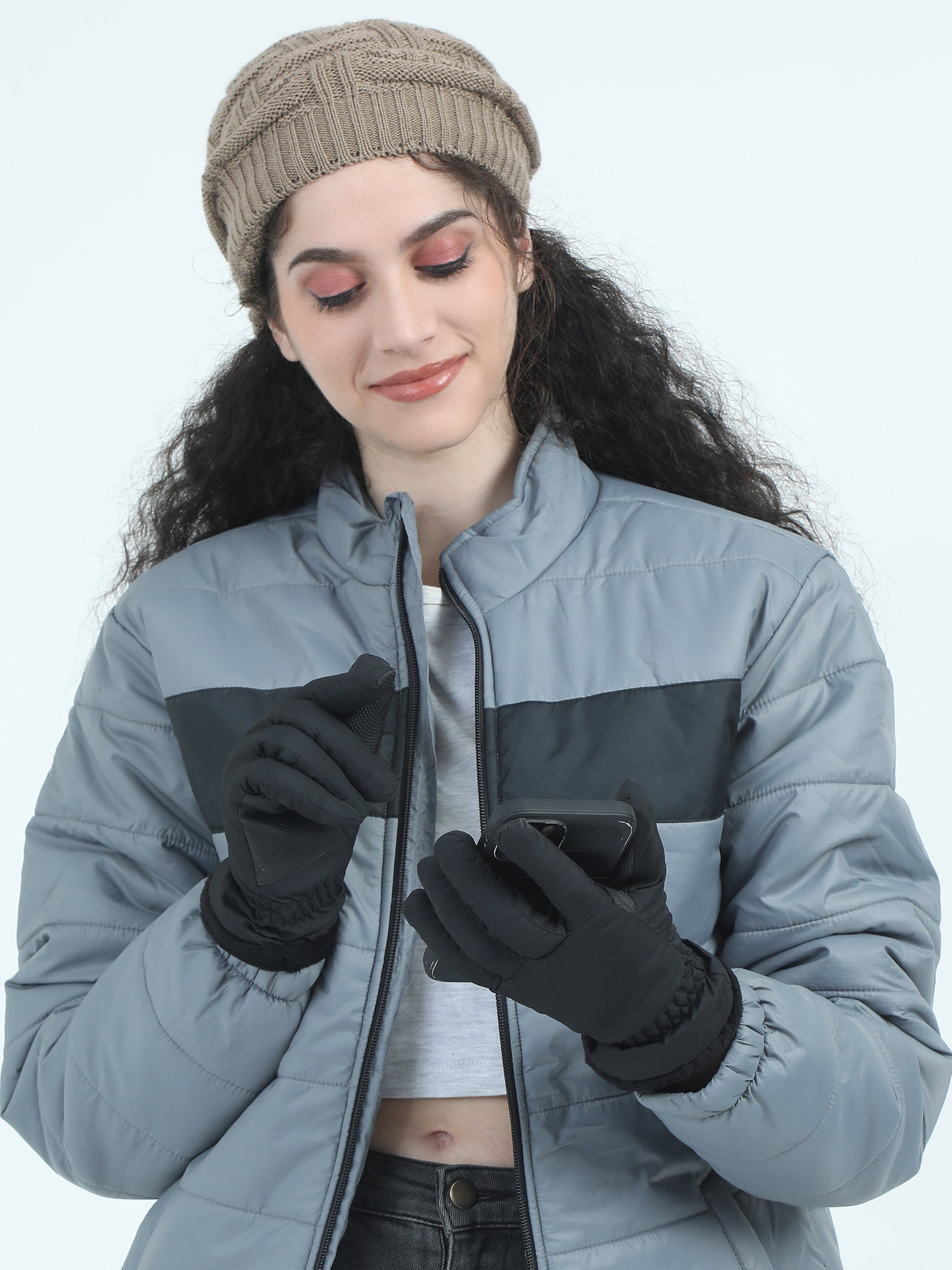 Woman wearing a gray winter jacket, black gloves, and a beige knit hat on a light blue background