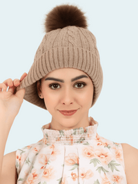 Woman wearing a Taupe knitted beanie with a brown pom-pom against a light blue background